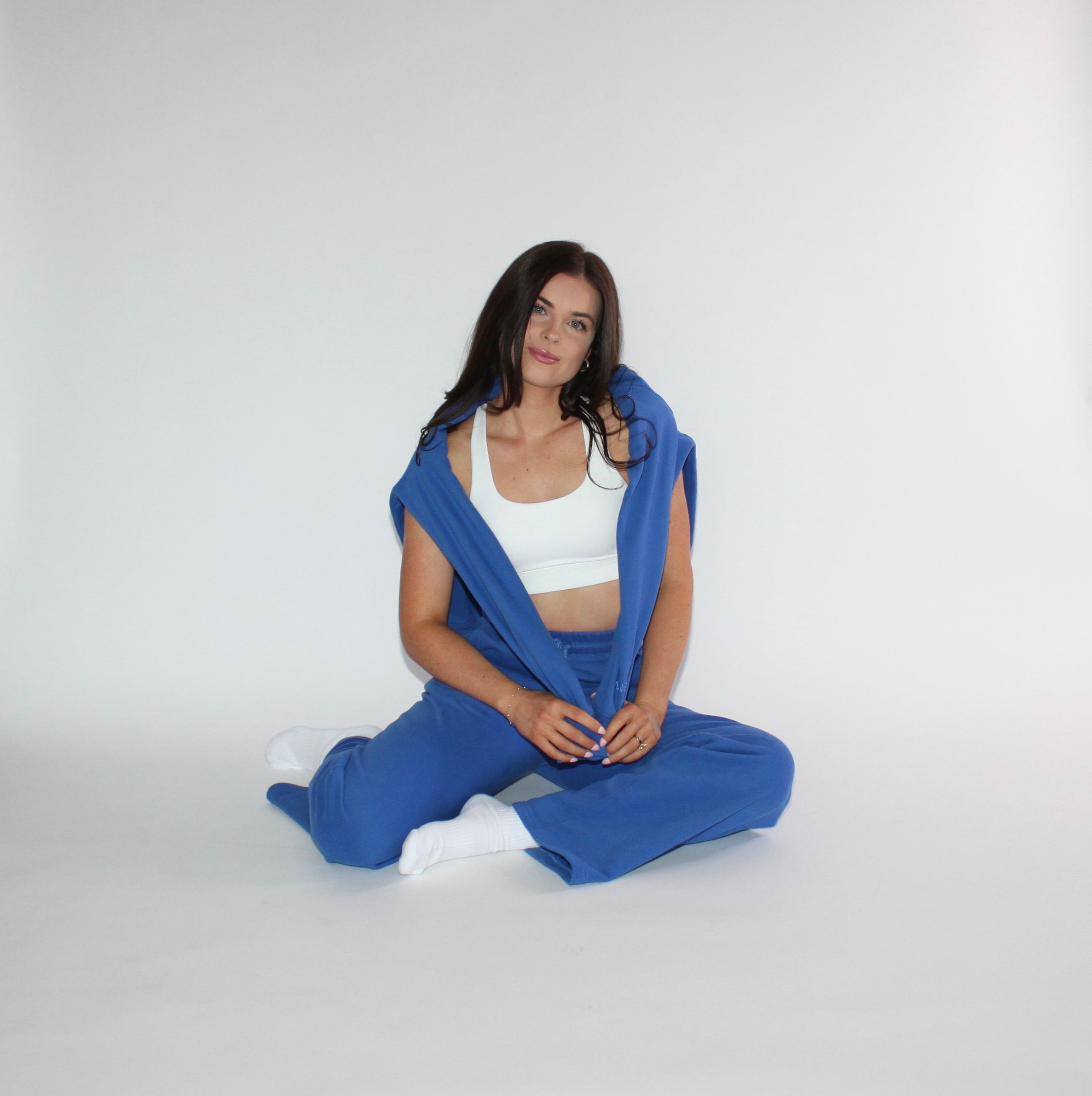A woman sits cross-legged on a light gray studio background wearing a matching royal blue lounge set, consisting of a sports bra and sweatpants, with a blue crewneck draped over her shoulders.
