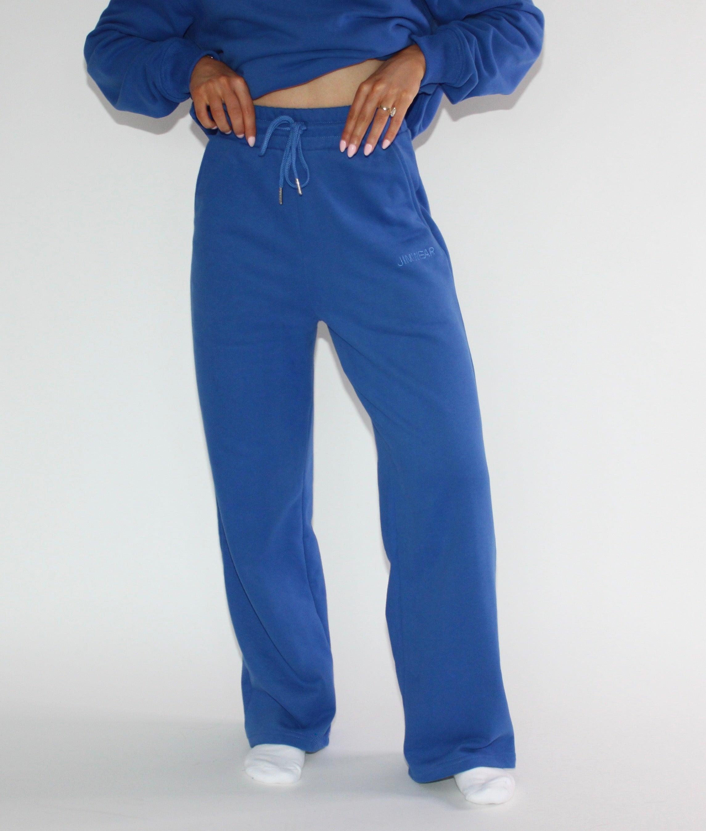 A person wearing a pair of wide-leg, relaxed-fit sweatpants in a medium blue color, paired with a matching blue cropped sweatshirt and white socks, against a plain light gray studio background.